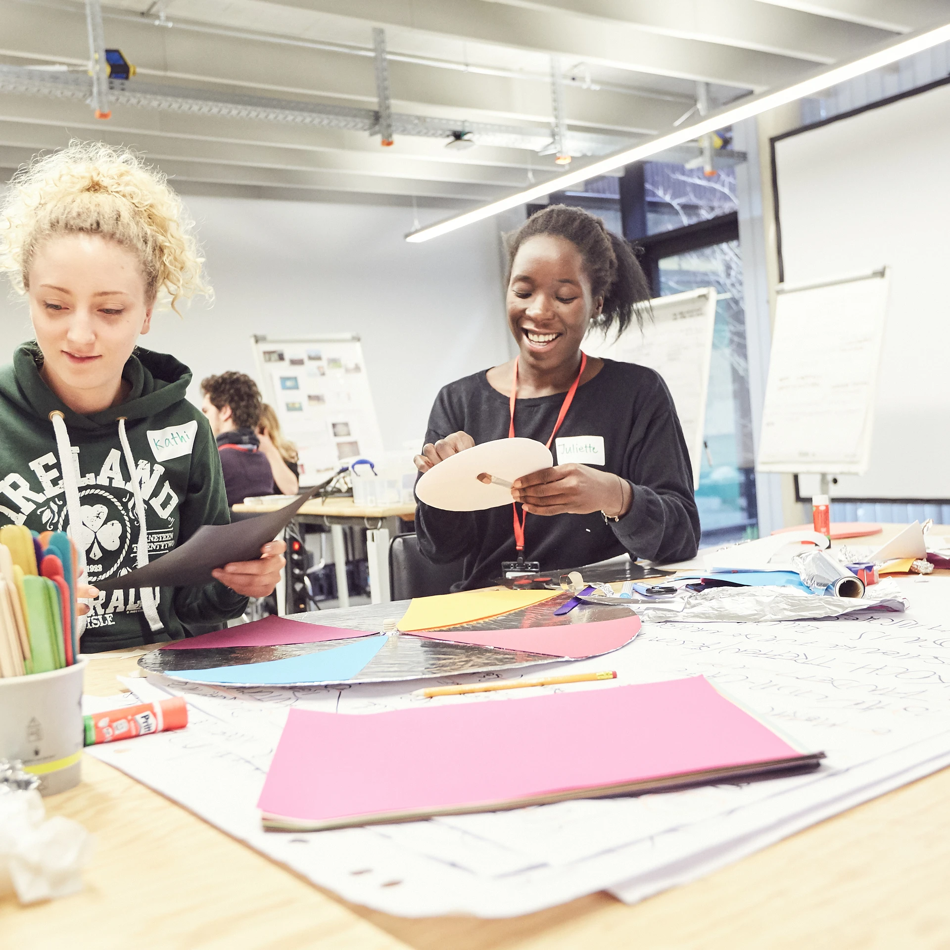 Zwei Frauen sitzen zusammen an einem Tisch und basteln zusammen eine Scheibe aus Pappe und Tonpapier. Auf dem Tisch ist Material zum Basteln verteilt.